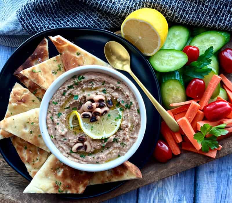 white bowl of black eyed pea hummus garnished with lemon slice, serve dwith bright fresh vegetables and pita