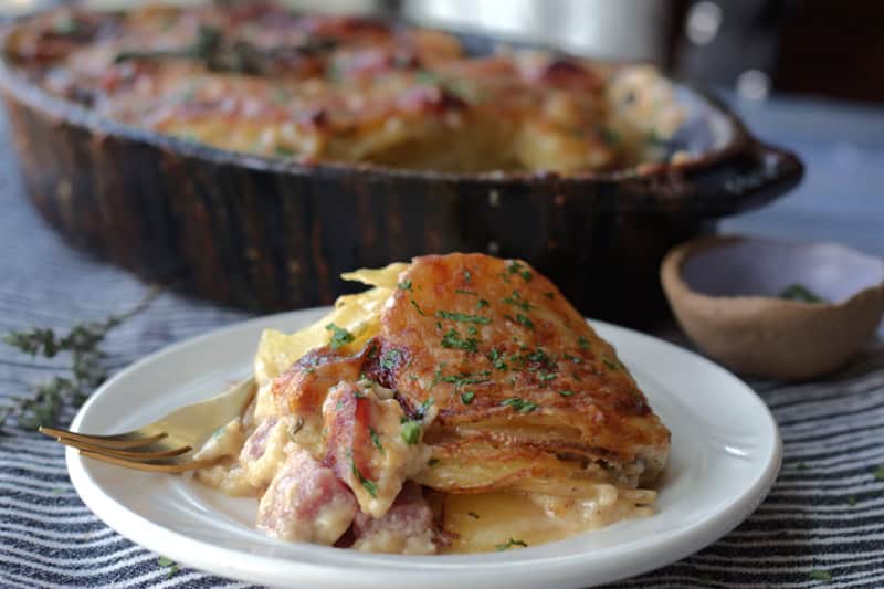 white plate with serving of scalloped potatoes and ham. black casserole dish in the background