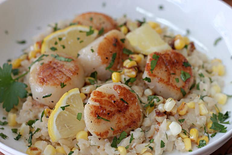 shallow white bowl with risotto and pan seared scallops garnished with parsley and lemon slices