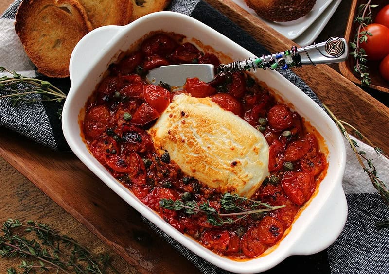 white baking dish with baked tomato goat cheese dip garnished with fresh thyme sprig