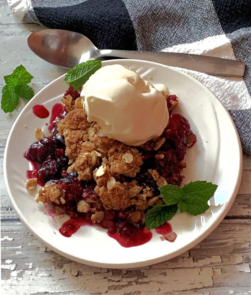 white plate with triple berry crisp topped with vanilla ice cream and garnished with mint leaves