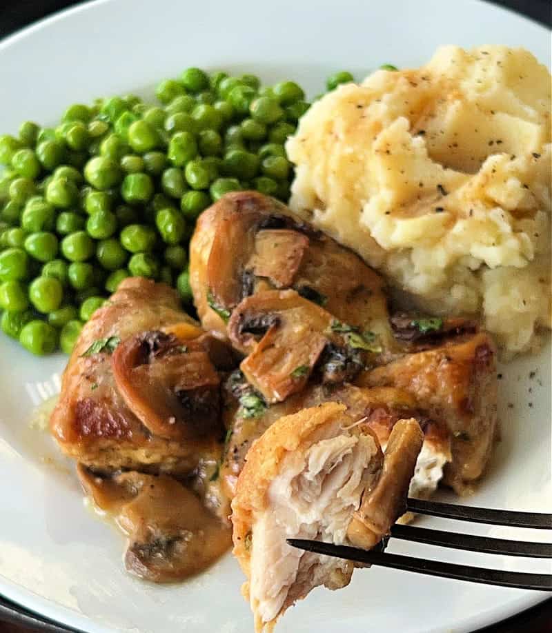 white plate with cheesy baked chicken, mashed potatoes and green peas. A fork is holding up a bite of the chicken