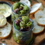 clear jars with marinated olives on a board with sliced baguette