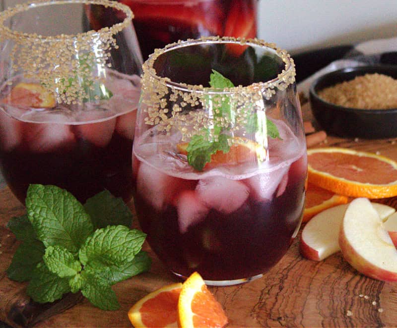 two glasses of red Thanksgiving Sangria in sugar rimmed glasses with sliced fruit on the side.