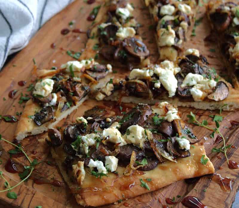 wooden board with mushroom goat cheese flatbread cut in squares, drizzled with balsamic vinegar