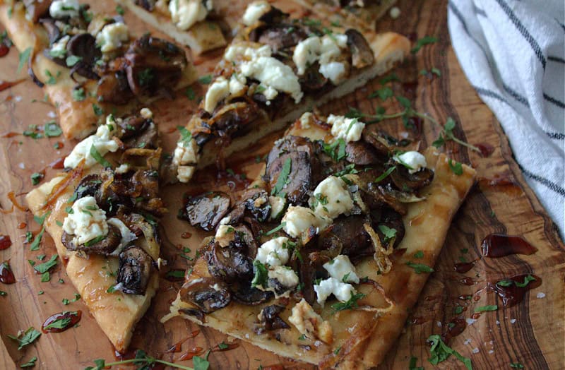 sliced mushroom goat cheese flatbread on a cutting board drizzled with balsamic glaze.
