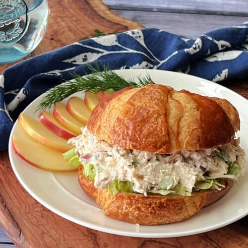 white plate with classic chicken salad on croissant with sliced apples on the side