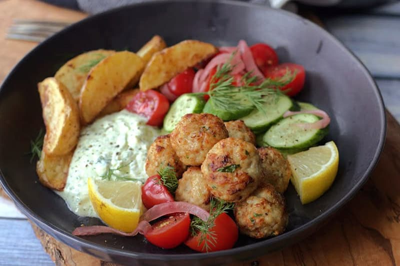 black bowl with air fryer meatballs, sliced cucumber and tomatoes and tzatziki dip.