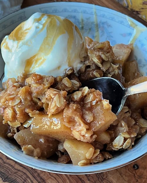 bowl of apple crisp with a scoop of vanilla ice cream and caramel topping and spoon scooping out some