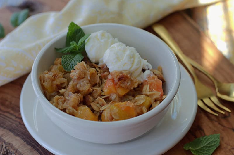 white bowl with peach crisp, scoops of vanilla ice cream and 2 small mint leaves for garnish