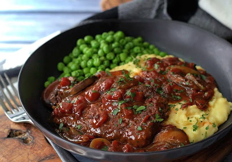 shallow black bowl with swiss steak, mashed potatoes and gravy and green peas