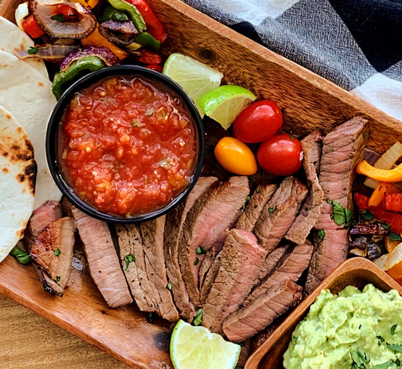 wooden board with sliced steak, guacamole, salsa, tortillas and grilled vegetables