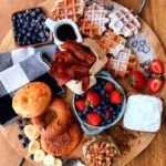 overhead view large board with breakfast foods - waffles, syrup, bagels, fruit, bacon, more