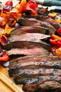 sliced flank steak on a cutting board surrounded by colorful grilled bell peppers