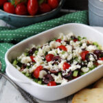shallow white dish with greek layered dip and container of pita bread in background