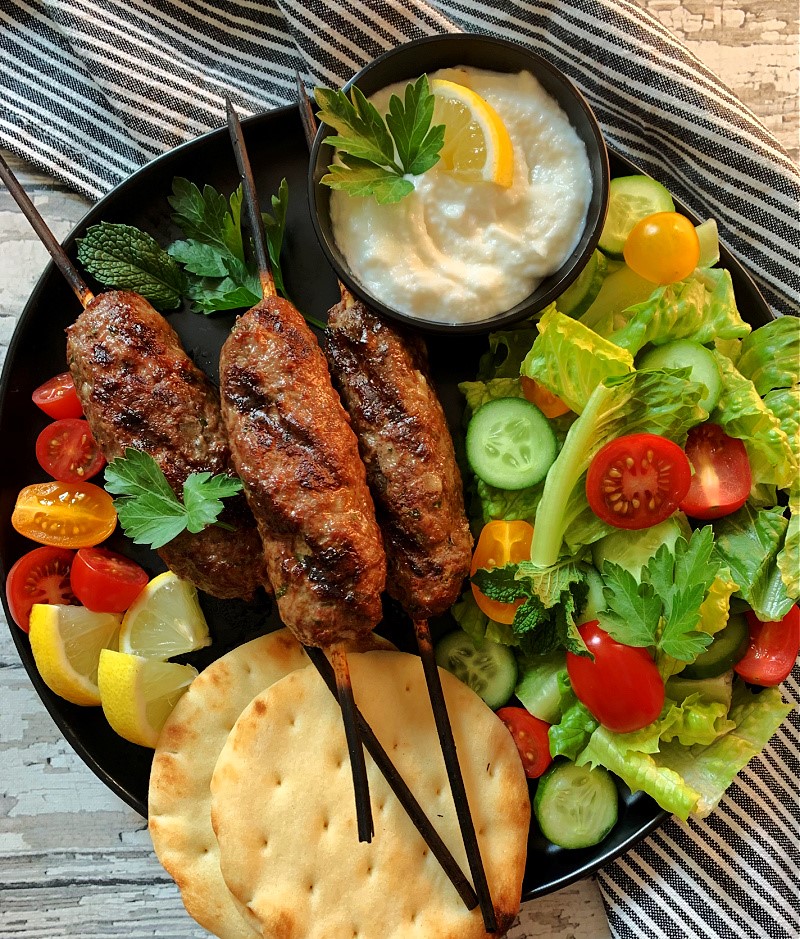black plate with 3 kofta kabobs, side salad, naan bread and a small bowl of garlic sauce