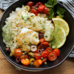 black bowl with rice, mediterranean baked fish and vegetables garnished with lemon slices