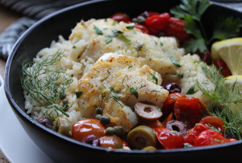 black plate with mediterranean baked fish, rice, tomatoes, capers and olives garnished with dill