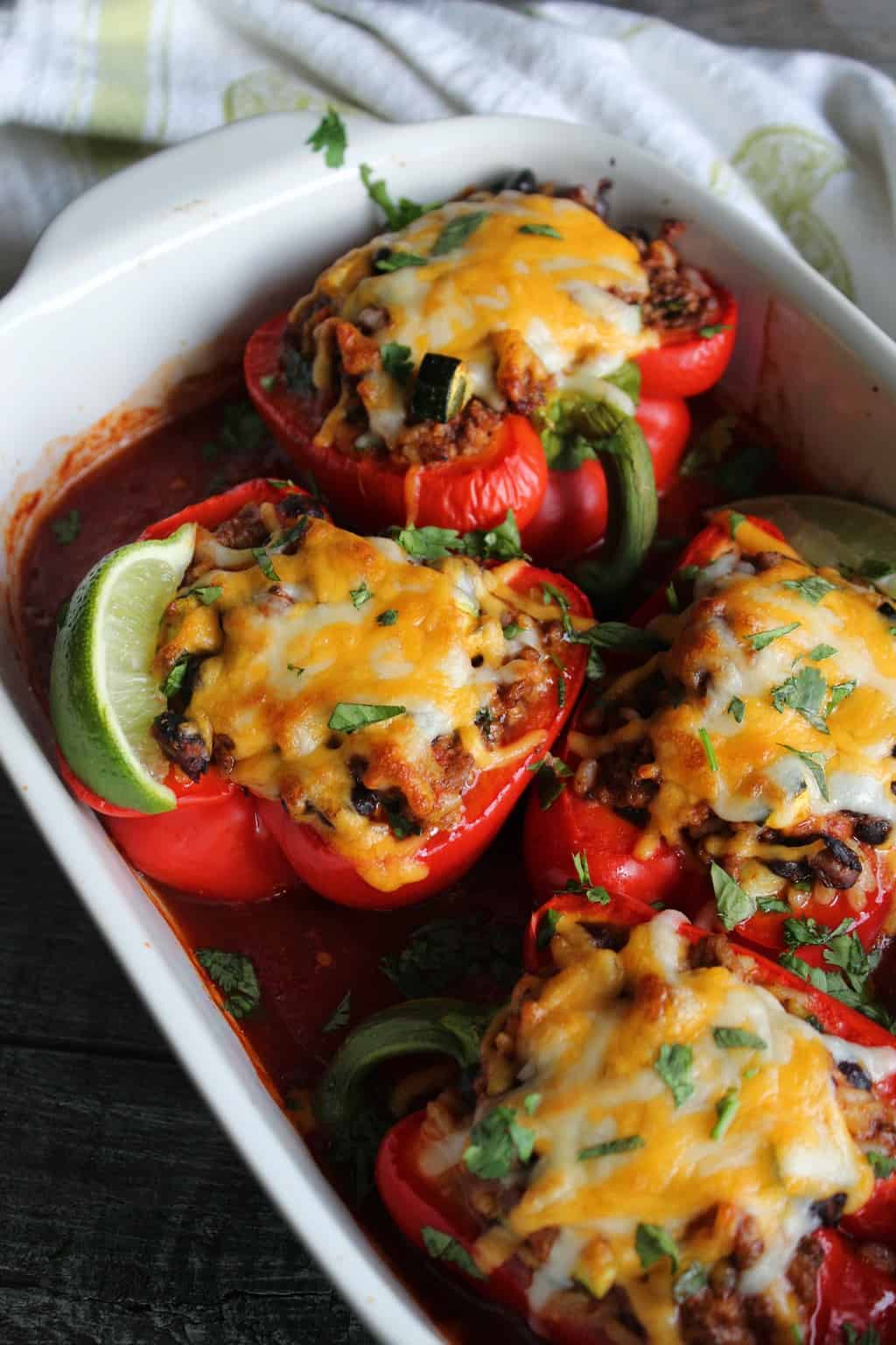 White casserole dish with stuffed red bell peppers in enchilada sauce topped with melted cheese