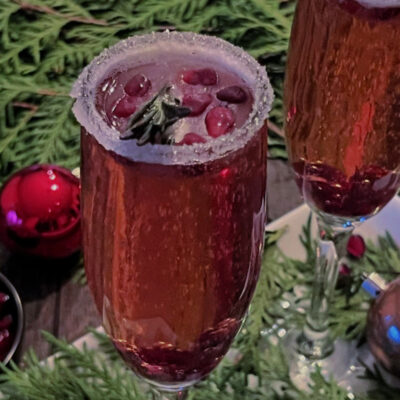 Two Mistletoe Mimosas garnished with rosemary and pomegranate seeds surrounded by greenery
