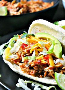 slow cooker salsa chicken ~ 4 ingredients, 5 minute prep for delicious, juicy, bold flavored chicken. Shown in soft shell taco topped with shredded lettuce, tomato, cheddar cheese and sliced avocado.